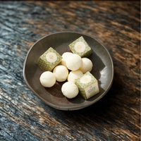 White bath melt and two green patterned wrapped boxes presented in a dark ceramic bowl on a rustic dark wooden surface.