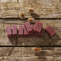 Several rectangular bars of dusty rose pink soap arranged on a rustic wooden surface, with dried thistle flowers placed around them.