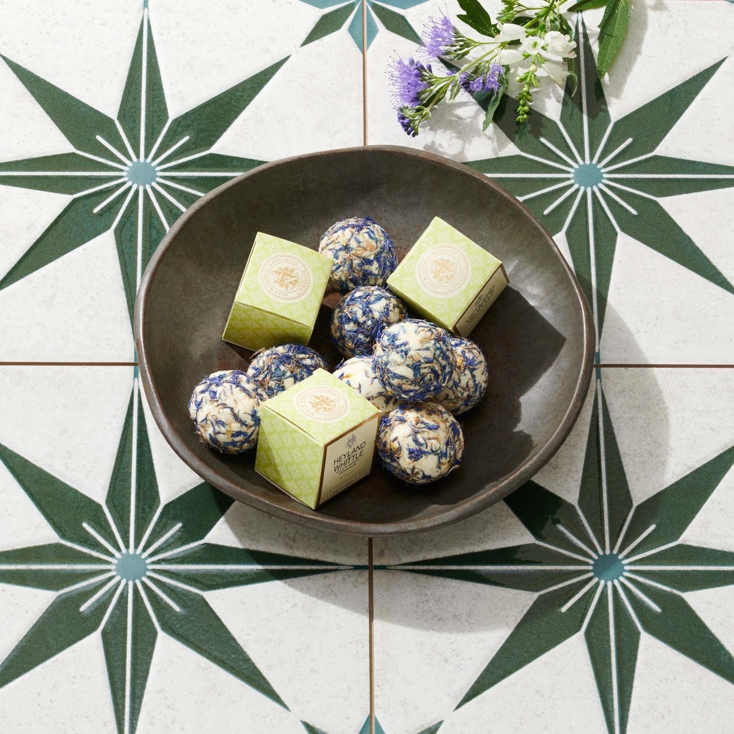 A dark brown ceramic bowl filled with round blue-and-white patterned bath melt and three light green square boxes, arranged on a white tile surface with a green starburst pattern and small purple flowers.