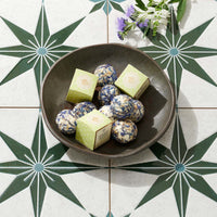 A dark brown ceramic bowl filled with round blue-and-white patterned bath melt and three light green square boxes, arranged on a white tile surface with a green starburst pattern and small purple flowers.