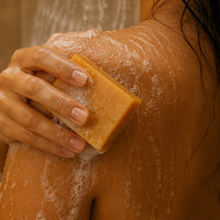 A close-up of a person's shoulder and back being washed with white lather while holding a rectangular bar of warm yellow soap.