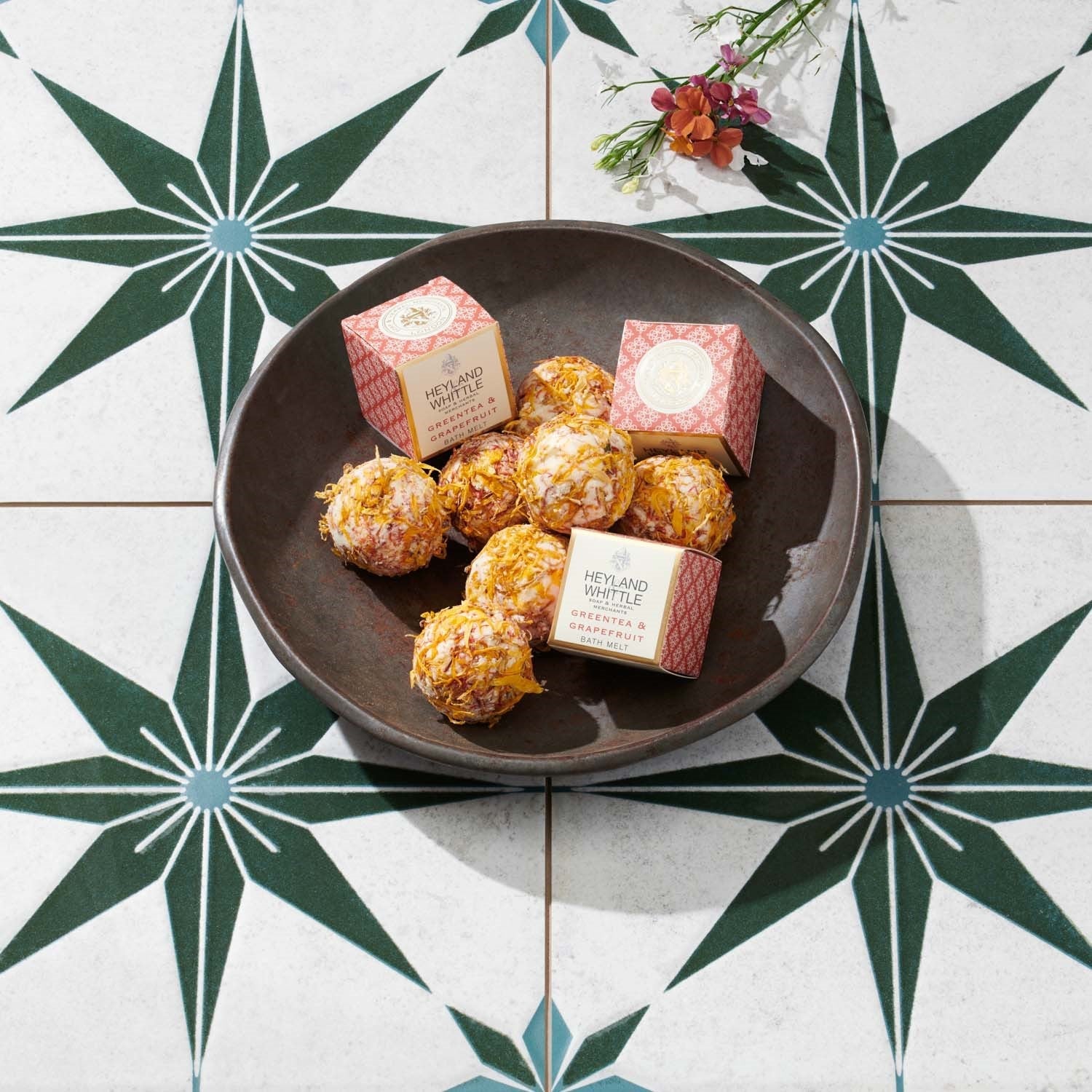 A dark brown ceramic plate holding several bath melts and two pink patterned boxes, photographed from above on a white tile surface with a green geometric star pattern and small flowers in the corner. 