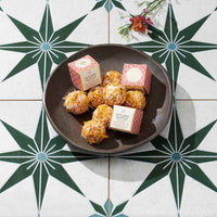 A dark brown ceramic plate holding several bath melts and two pink patterned boxes, photographed from above on a white tile surface with a green geometric star pattern and small flowers in the corner. 